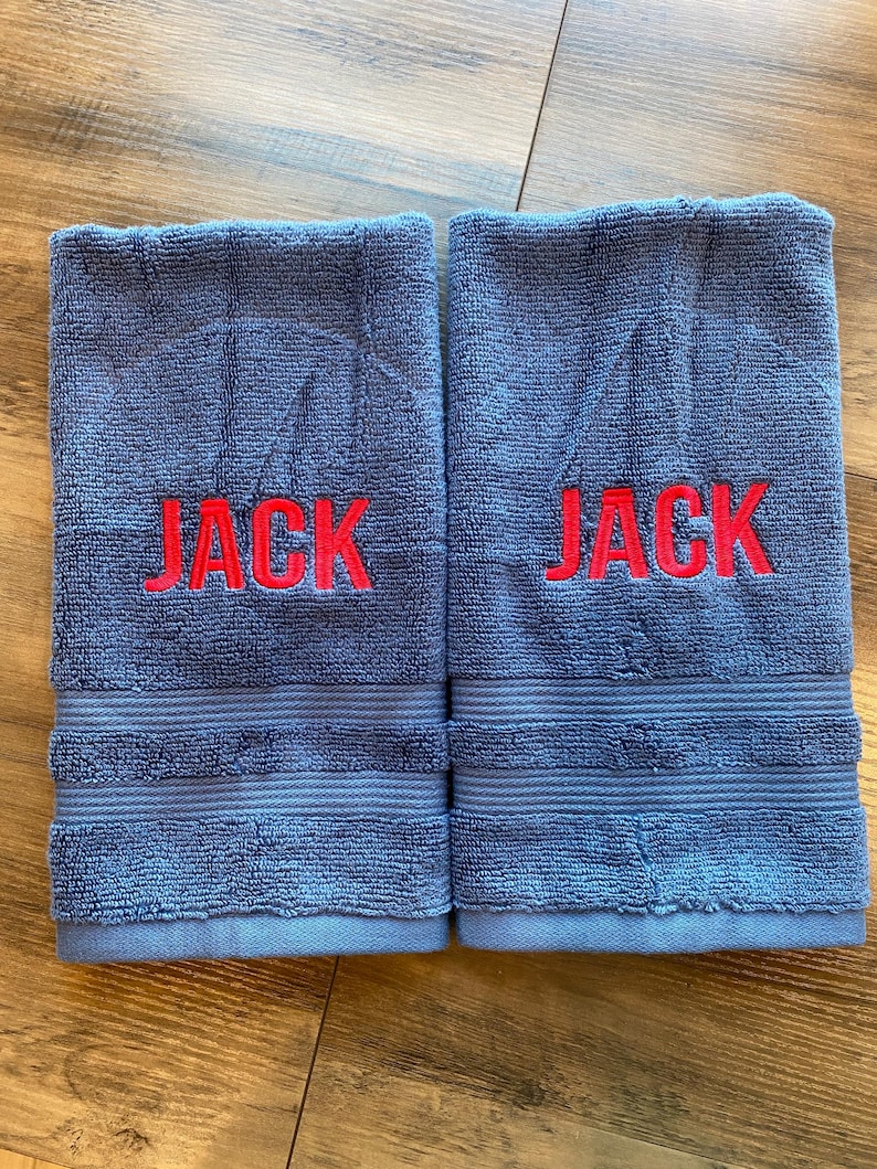 May include: Two rectangular blue terry cloth towels featuring the name JACK embroidered in bold red capital letters across the centre. Each towel has a textured surface with horizontal ribbed bands near the bottom edge and finished hems.