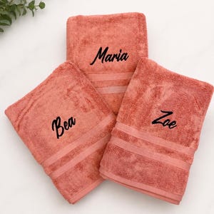 May include: Three folded coral-coloured cotton towels, each featuring a distinct name embroidered in black script lettering. The names Maria, Bea, and Zoe are centred on the front of each individual towel, which also includes two horizontal bands.