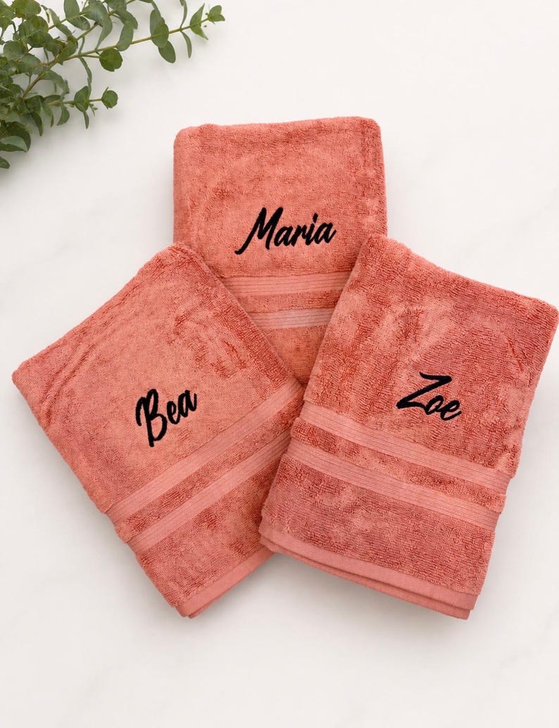 May include: Three folded coral-coloured cotton towels, each featuring a distinct name embroidered in black script lettering. The names Maria, Bea, and Zoe are centred on the front of each individual towel, which also includes two horizontal bands.