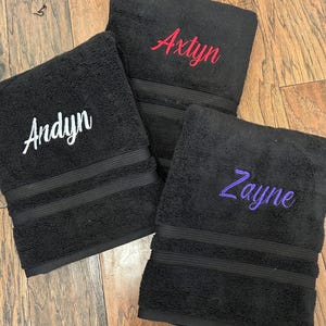May include: Three black rectangular towels featuring embroidered names in cursive script. One towel displays the name Andyn in white, another shows Axtyn in red, and the third features Zayne in purple. Each towel has a textured surface and horizontal bands.