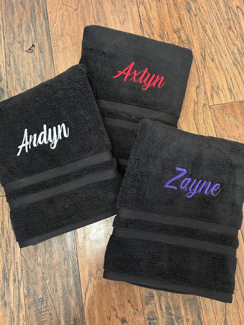 May include: Three black rectangular towels featuring embroidered names in cursive script. One towel displays the name Andyn in white, another shows Axtyn in red, and the third features Zayne in purple. Each towel has a textured surface and horizontal bands.