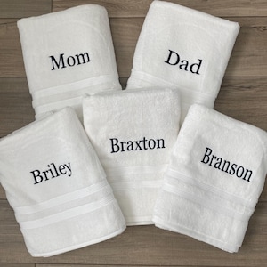 May include: Five white rectangular bath towels, each featuring dark navy blue embroidered text. The towels are personalised with the names Mom, Dad, Braxton, Briley, and Branson, with each towel displaying a single name in a serif font style.