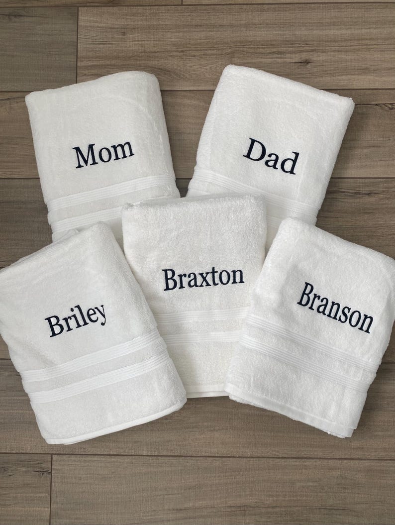 May include: Five white rectangular bath towels, each featuring dark navy blue embroidered text. The towels are personalised with the names Mom, Dad, Braxton, Briley, and Branson, with each towel displaying a single name in a serif font style.