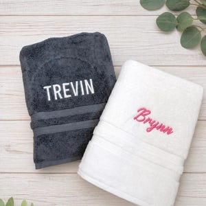 May include: A dark grey towel with the name TREVIN embroidered in white block letters and a white towel with the name Brynn embroidered in pink cursive script. Both towels feature a horizontal ribbed texture detail near the bottom edge.