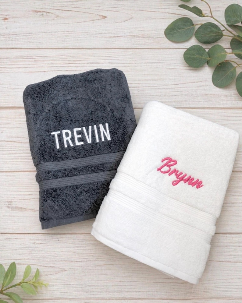 May include: A dark grey towel with the name TREVIN embroidered in white block letters and a white towel with the name Brynn embroidered in pink cursive script. Both towels feature a horizontal ribbed texture detail near the bottom edge.