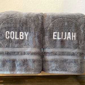May include: Two folded charcoal grey bath towels featuring white embroidered text. The left towel displays the name COLBY, and the right towel displays the name ELIJAH. Each towel has a horizontal ribbed texture band above the bottom hem.