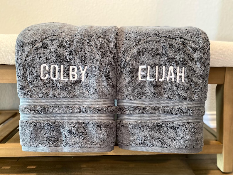 May include: Two folded charcoal grey bath towels featuring white embroidered text. The left towel displays the name COLBY, and the right towel displays the name ELIJAH. Each towel has a horizontal ribbed texture band above the bottom hem.