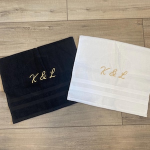 May include: A black towel and a white towel, each featuring the embroidered gold text K & L. Both towels have a textured fabric surface and two horizontal decorative bands near the bottom edge, providing a clean and simple aesthetic design.