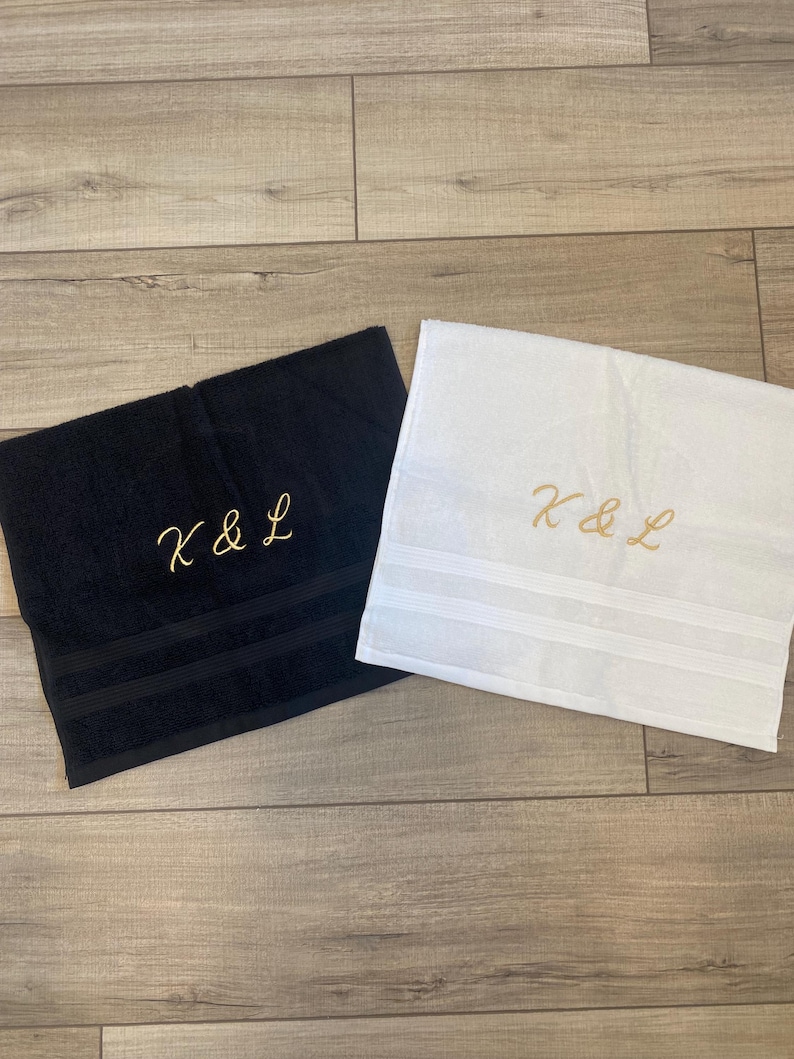 May include: A black towel and a white towel, each featuring the embroidered gold text K & L. Both towels have a textured fabric surface and two horizontal decorative bands near the bottom edge, providing a clean and simple aesthetic design.