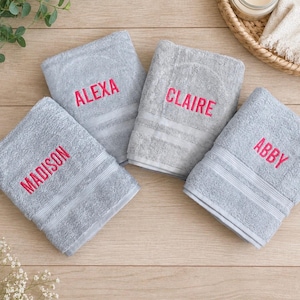 May include: Four folded light grey towels featuring embroidered names in bright pink thread. The names Madison, Alexa, Claire, and Abby are stitched onto the fabric. Each towel has a textured surface with two horizontal bands near the bottom edge.