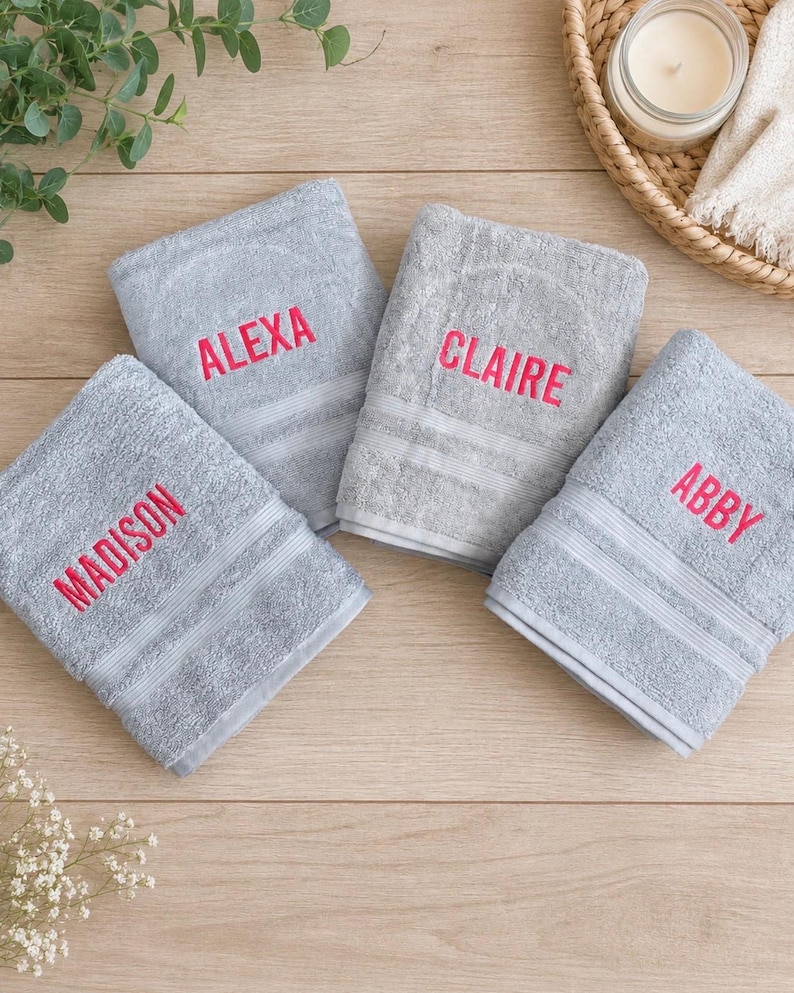 May include: Four folded light grey towels featuring embroidered names in bright pink thread. The names Madison, Alexa, Claire, and Abby are stitched onto the fabric. Each towel has a textured surface with two horizontal bands near the bottom edge.