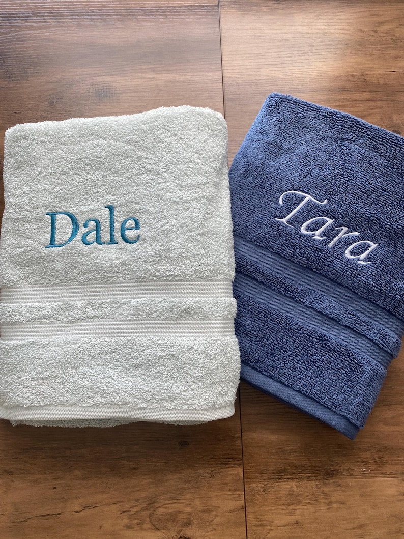 May include: A white bath towel embroidered with the name Dale in blue thread and a blue bath towel embroidered with the name Tara in white thread. Both towels feature a textured terry cloth surface and a horizontal ribbed band detail near the bottom.