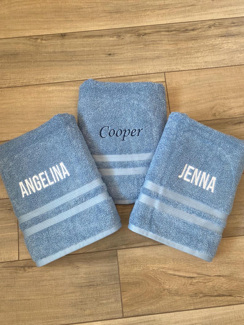 May include: Three light blue bath towels featuring embroidered names. The left towel displays the name ANGELINA in white, the right towel displays JENNA in white, and the centre towel displays the name Cooper in dark blue embroidery.