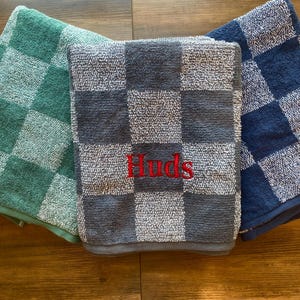 May include: Three folded checkered towels in shades of green, gray, and blue. The center towel is gray with the name "Huds" embroidered in red. The towels are arranged on a wooden surface.