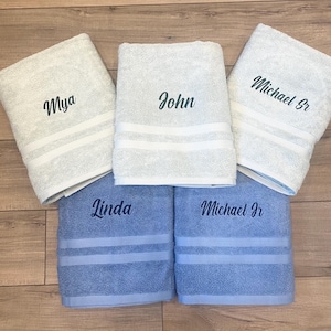 May include: Five folded bath towels featuring embroidered names. Three towels are light grey with the names Mya, John, and Michael Sr. Two towels are light blue with the names Linda and Michael Jr. Each towel has two horizontal white stripes.