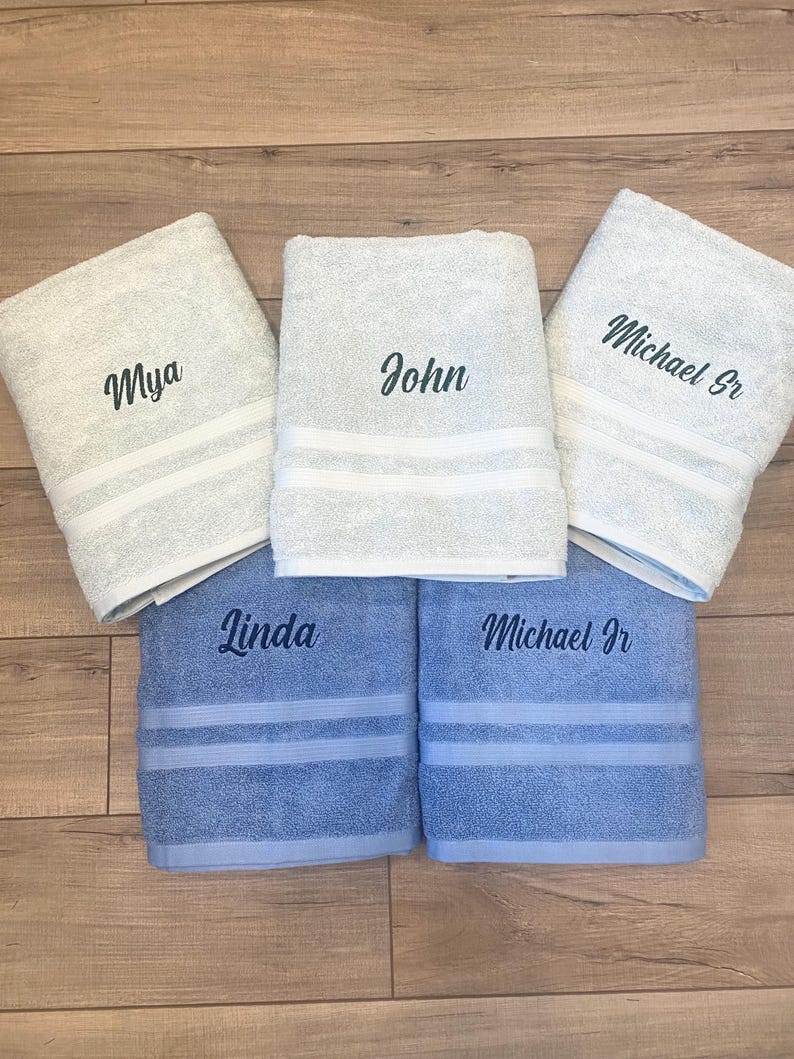 May include: Five folded bath towels featuring embroidered names. Three towels are light grey with the names Mya, John, and Michael Sr. Two towels are light blue with the names Linda and Michael Jr. Each towel has two horizontal white stripes.