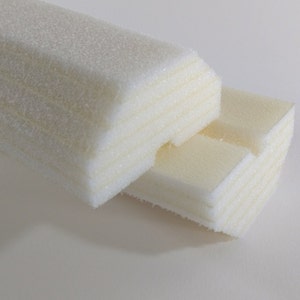 May include: Close-up of two rectangular, off-white foam blocks. The blocks are layered, with visible horizontal lines. The foam has a slightly textured surface and a soft appearance. The blocks are positioned against a plain white background.