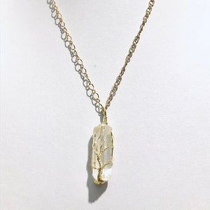 Op de afbeelding: Een gouden ketting met een hanger van helder kwartskristal met een draadgewikkeld boomontwerp.