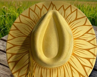 May include: A yellow fedora-style hat with a sunflower design on the brim. The hat has a matching yellow tassel and is set against a natural outdoor background. The hat is made of a soft material.