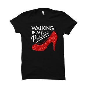 Puede incluir: Camiseta negra con el texto "Walking in my Purpose" y un zapato de tacón alto rojo con purpurina.
