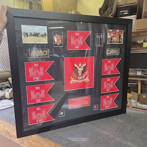 May include: A framed display featuring military insignia and photographs. The display includes red and white pennants with the number "20" and various unit designations. The frame is black and contains multiple images and emblems.