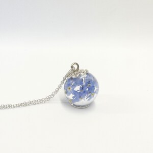 Peut inclure: Un collier argenté avec un pendentif sphérique transparent rempli de petites fleurs bleu clair. Le pendentif est suspendu à une fine chaîne argentée. Les fleurs sont visibles à l'intérieur de la sphère.