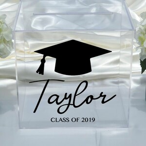 May include: Clear acrylic graduation box with a slot on top. A black graduation cap graphic and the name "Taylor" are displayed, along with "CLASS OF 2019" in black script. Ideal for graduation parties.