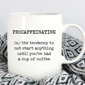 May include: White ceramic coffee mug with black text that reads "PROCAFFEINATING (n.) the tendency to not start anything until you've had a cup of coffee"