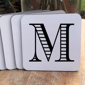 May include: Set of four white square coasters with black letter M printed on the top. The letter M has a striped pattern.