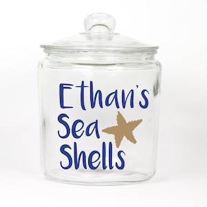 May include: A clear glass jar with a lid, decorated with a blue and gold design. The jar says "Ethan's Sea Shells" with a gold starfish.