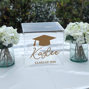 May include: Clear acrylic graduation box with a slot on top. The box features a gold mortarboard graphic and the name "Kaylee" with "CLASS OF 2020" in a script font. White flowers in glass vases flank the box.