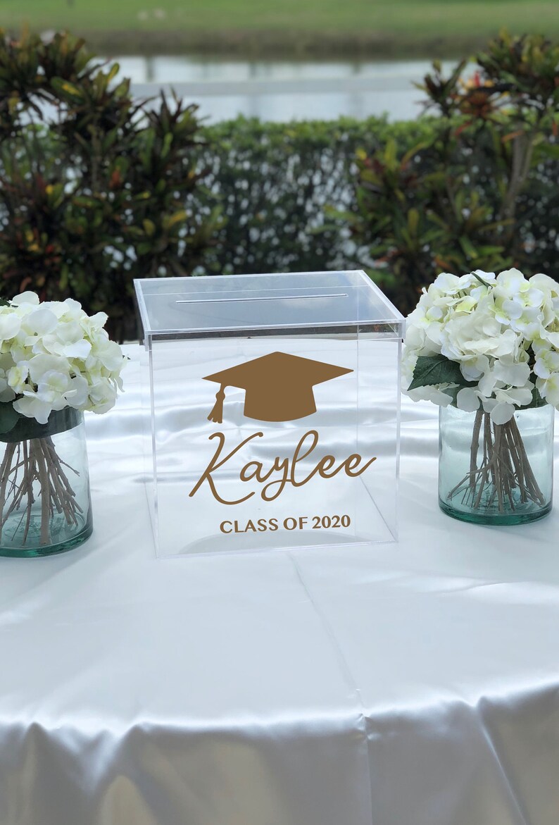May include: Clear acrylic graduation box with a slot on top. The box features a gold mortarboard graphic and the name "Kaylee" with "CLASS OF 2020" in a script font. White flowers in glass vases flank the box.