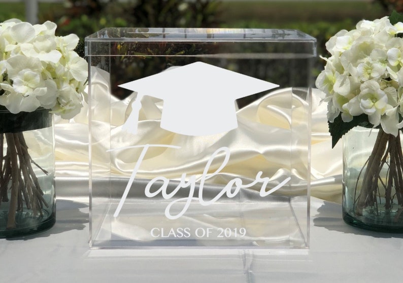 May include: A clear acrylic graduation box with a white mortarboard design and the name "Taylor" in script. The text "CLASS OF 2019" is below the name. White flowers in glass vases flank the box.