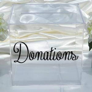 May include: Clear acrylic donation box with a slot on top. The word "Donations" is written in black script across the front of the box. The box is sitting on a white surface.