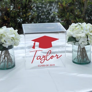 May include: Clear acrylic box with a red graduation cap graphic and the name "Taylor" with "CLASS OF 2019" in red script. The box is on a white tablecloth, flanked by vases of white flowers.