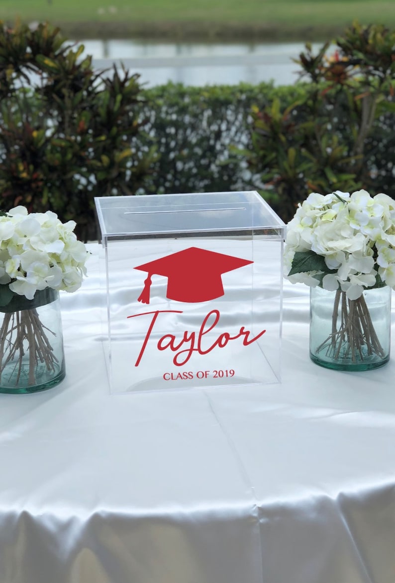 May include: Clear acrylic box with a red graduation cap graphic and the name "Taylor" with "CLASS OF 2019" in red script. The box is on a white tablecloth, flanked by vases of white flowers.