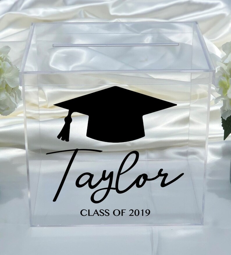 May include: Clear acrylic graduation box with a slot on top. A black graduation cap graphic and the name "Taylor" are displayed, along with "CLASS OF 2019" in black script. Ideal for graduation parties.