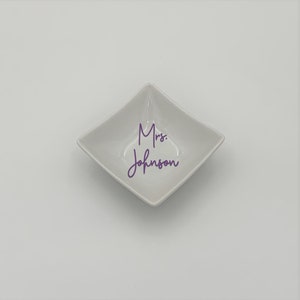 May include: A white square dish with rounded edges. The dish has the text "Mrs. Johnson" written in purple cursive.