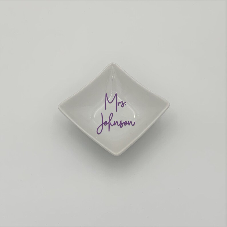 May include: A white square dish with rounded edges. The dish has the text "Mrs. Johnson" written in purple cursive.