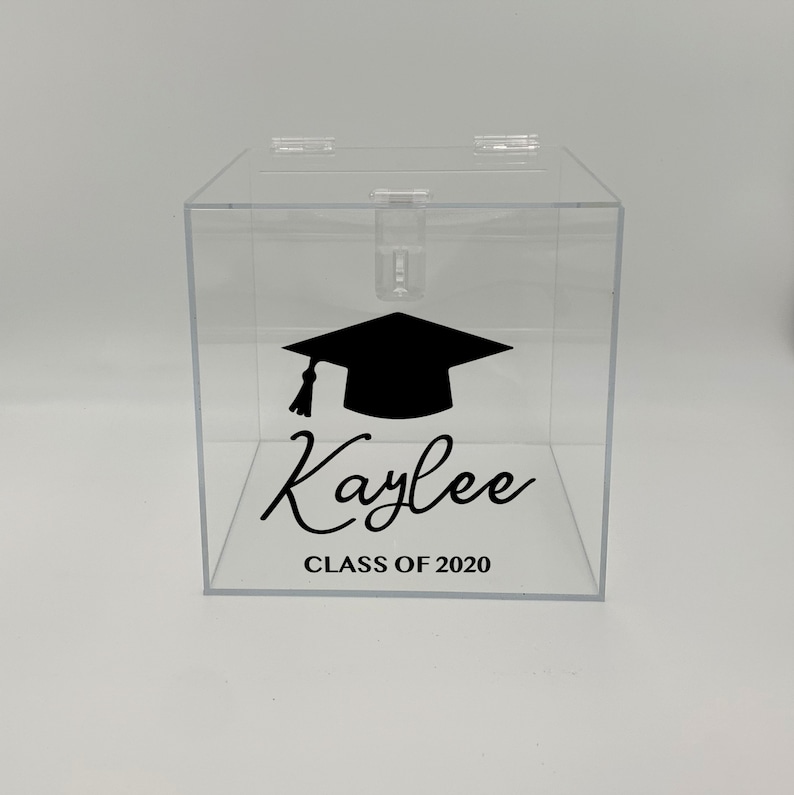 May include: Clear acrylic graduation box with a black mortarboard graphic and the name "Kaylee" in cursive. The text "CLASS OF 2020" is below the name. The box has a hinged lid and a slot for inserting items.