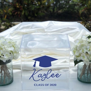 May include: Clear acrylic graduation card box with a blue mortarboard design and the name "Kaylee" and "Class of 2020" in blue script. The box is displayed on a white satin surface, flanked by white floral arrangements.