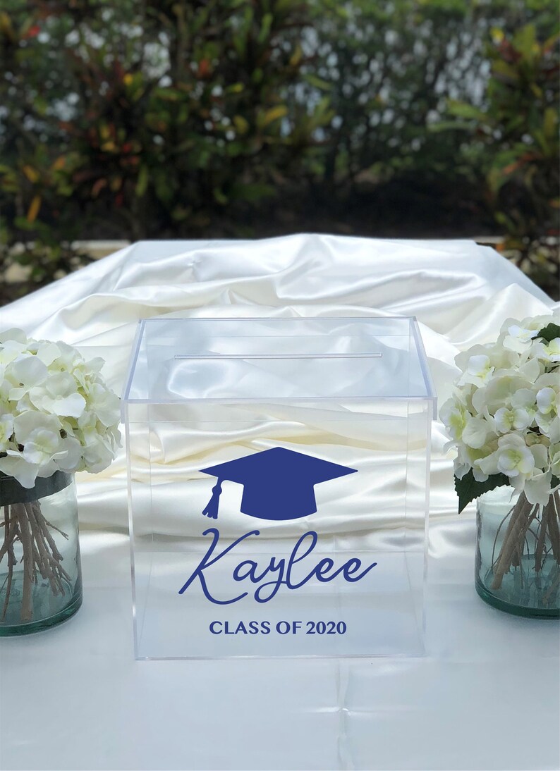 May include: Clear acrylic graduation card box with a blue mortarboard design and the name "Kaylee" and "Class of 2020" in blue script. The box is displayed on a white satin surface, flanked by white floral arrangements.