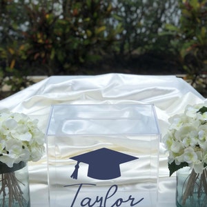 May include: Clear acrylic box with a navy blue graduation cap graphic and the name "Taylor" in script. The text "CLASS OF 2019" is below the name. White flowers in glass vases flank the box.