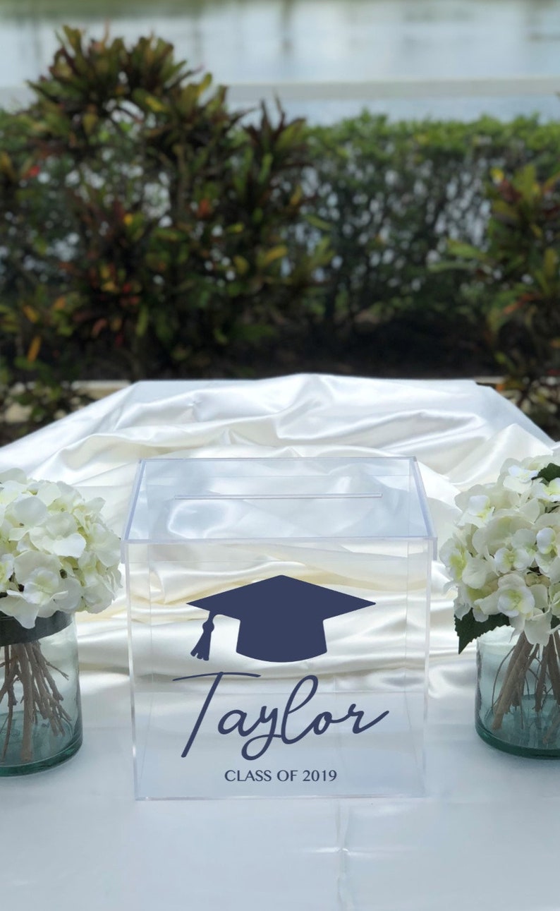 May include: Clear acrylic box with a navy blue graduation cap graphic and the name "Taylor" in script. The text "CLASS OF 2019" is below the name. White flowers in glass vases flank the box.
