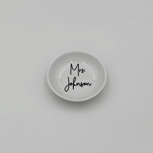 May include: White ceramic ring dish with the text "Mrs. Johnson" in black cursive lettering.
