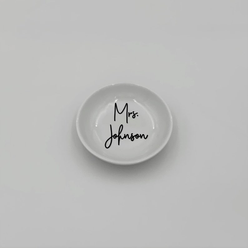 May include: White ceramic ring dish with the text "Mrs. Johnson" in black cursive lettering.