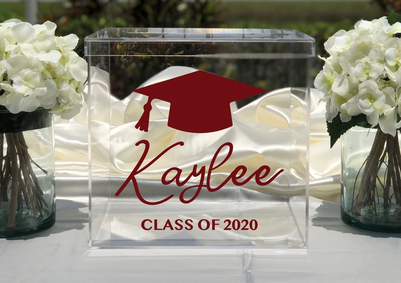 May include: Clear acrylic graduation box with a maroon mortarboard and the name "Kaylee" in cursive. The text "CLASS OF 2020" is below the name. The box is displayed with white flowers in the background.