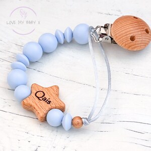 May include: A baby pacifier with a light blue beaded chain and a wooden star-shaped charm engraved with the name "Qais". The pacifier features a wooden clip and a round wooden piece for the nipple. The text "LOVE MY BABY X" is in the upper left corner.