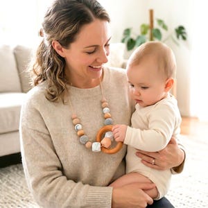 May include: A beige teething necklace with wooden and gray beads, and a wooden ring teether. The necklace is being held by a person holding a baby. The baby is wearing a white onesie.