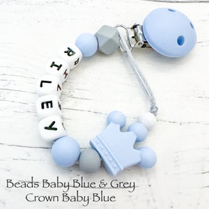 May include: A dummy clip featuring light blue and grey silicone beads, including white letter beads spelling the name RILEY. The clip includes a light blue crown-shaped bead and a circular light blue plastic clip attached by a grey cord.